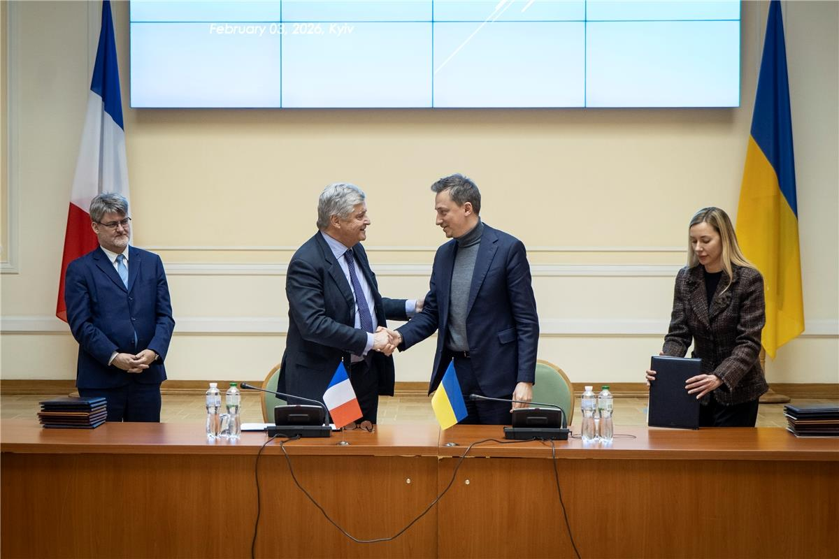 Двоє чоловіків потискають руки за столом із прапорцями Франції та України на фоні відповідних державних прапорів. Поруч стоять чоловік у костюмі та жінка з папкою в руках.