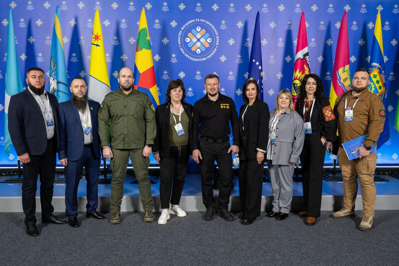 Група з дев'яти осіб стоїть у ряд на фоні синього банера з емблемою Конгресу місцевих та регіональних влад і низки прапорів. Присутні одягнені в офіційні костюми, повсякденний одяг та мілітарі-стиль; дехто має бейджі учасників заходу.