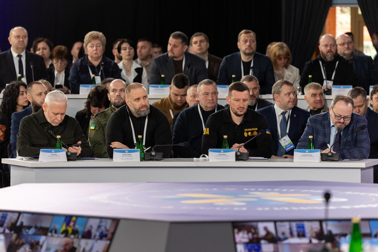 Велика група людей на засіданні: четверо чоловіків за столом із мікрофонами в першому ряду та кілька рядів учасників за ними. На передньому плані — екран із відеозв'язком.