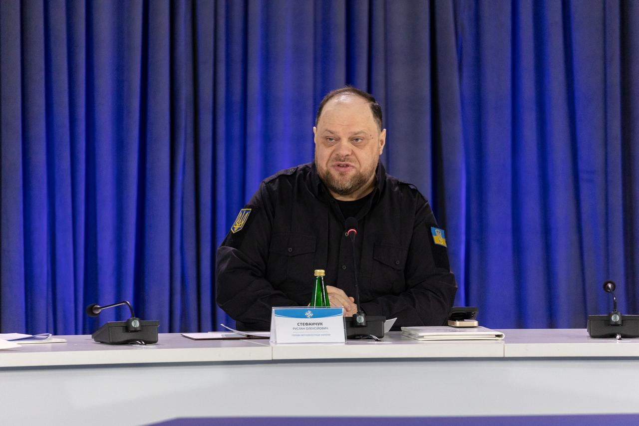 Чоловік у чорній тактичній сорочці з шевронами сидить за білим столом із мікрофонами та табличкою на фоні синіх штор. Перед ним стоять пляшка води, папка та телефон.