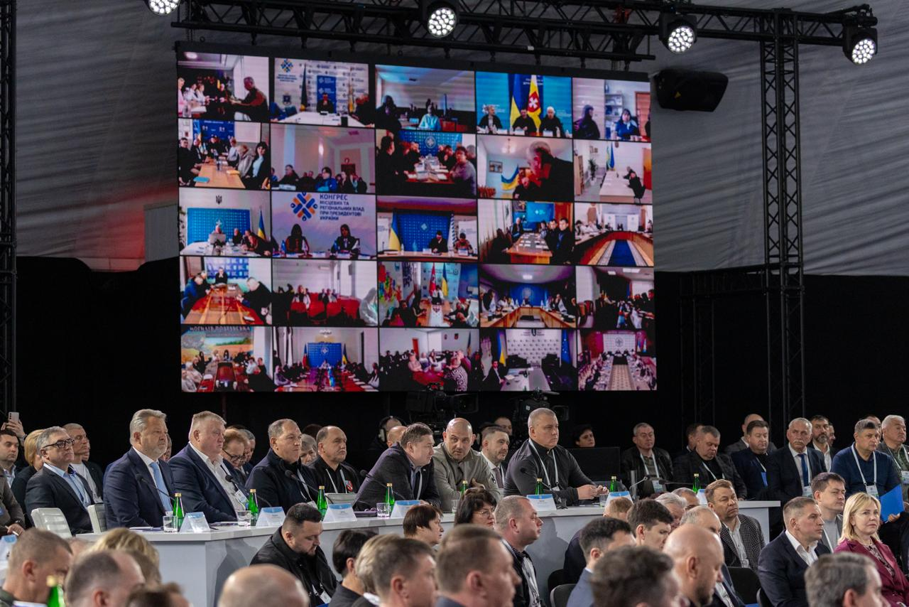 Масштабний форум у великому залі з великим екраном, на якому транслюється відеозв'язок із багатьма учасниками. На передньому плані за столами та в залі сидить численна аудиторія в офіційному та повсякденному вбранні.