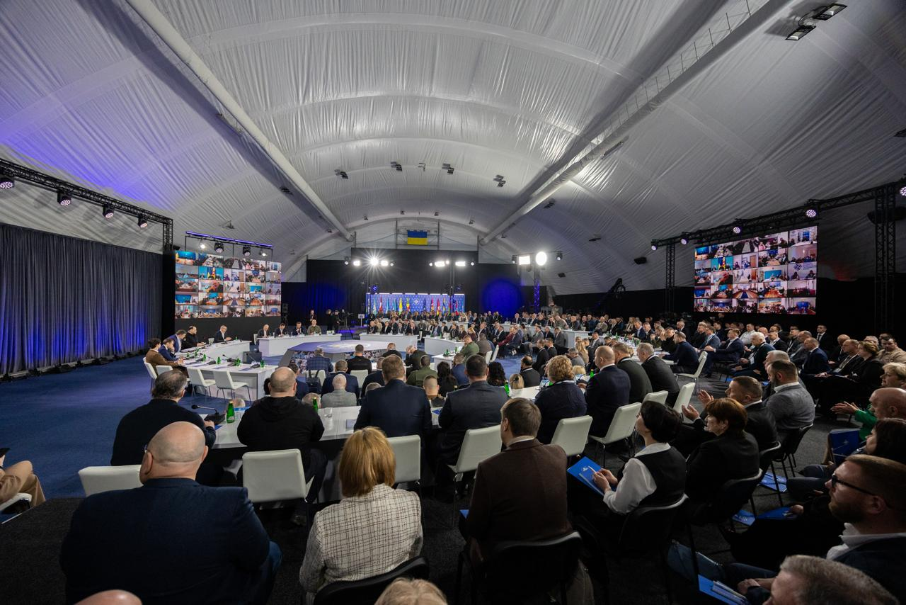 Масштабний форум у великому залі під тентовим дахом із численною аудиторією. По боках встановлено два великі екрани з вікнами відеозв'язку багатьох учасників. У центрі за круглим столом триває засідання на фоні прапорів України та Євросоюзу.