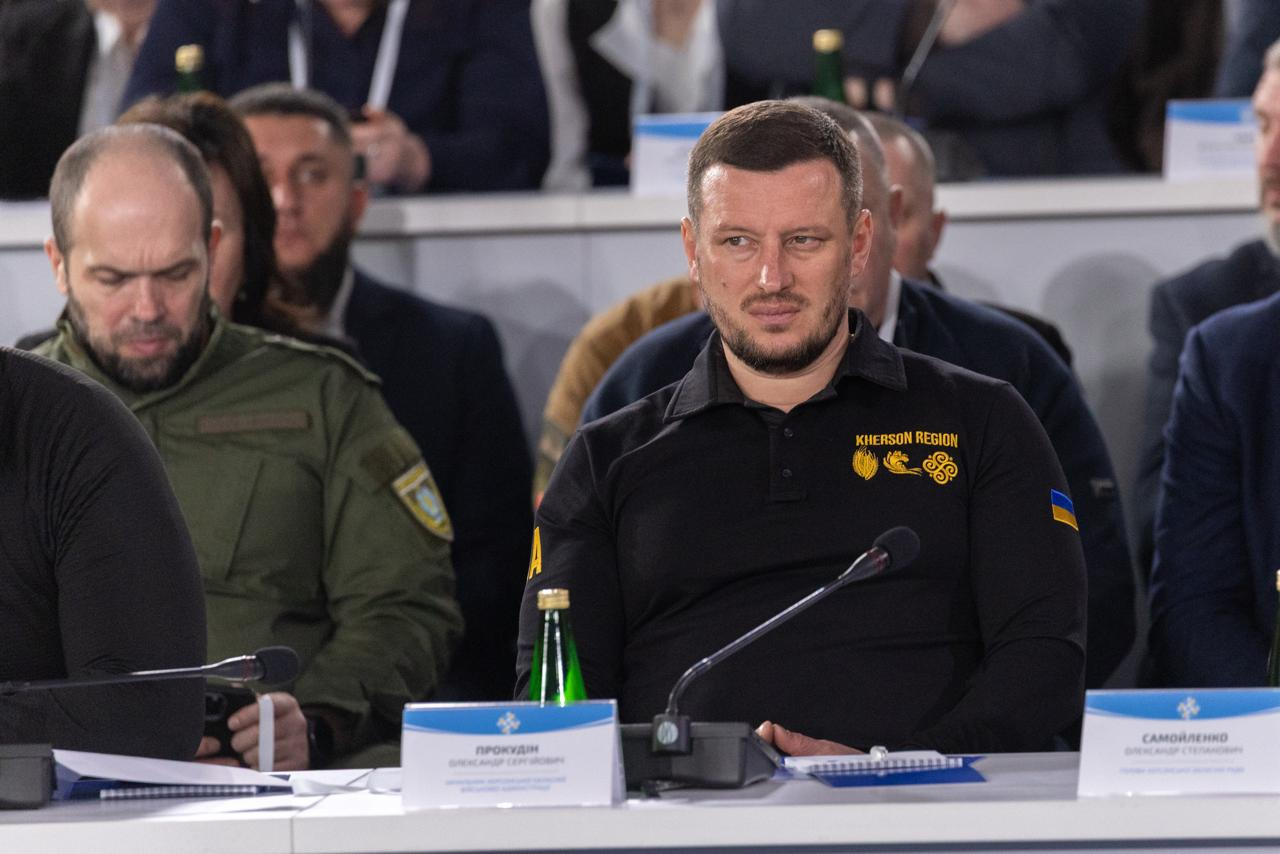 Чоловік у чорному поло з написом «KHERSON REGION» сидить за столом із мікрофонами та іменними табличками під час офіційного заходу.