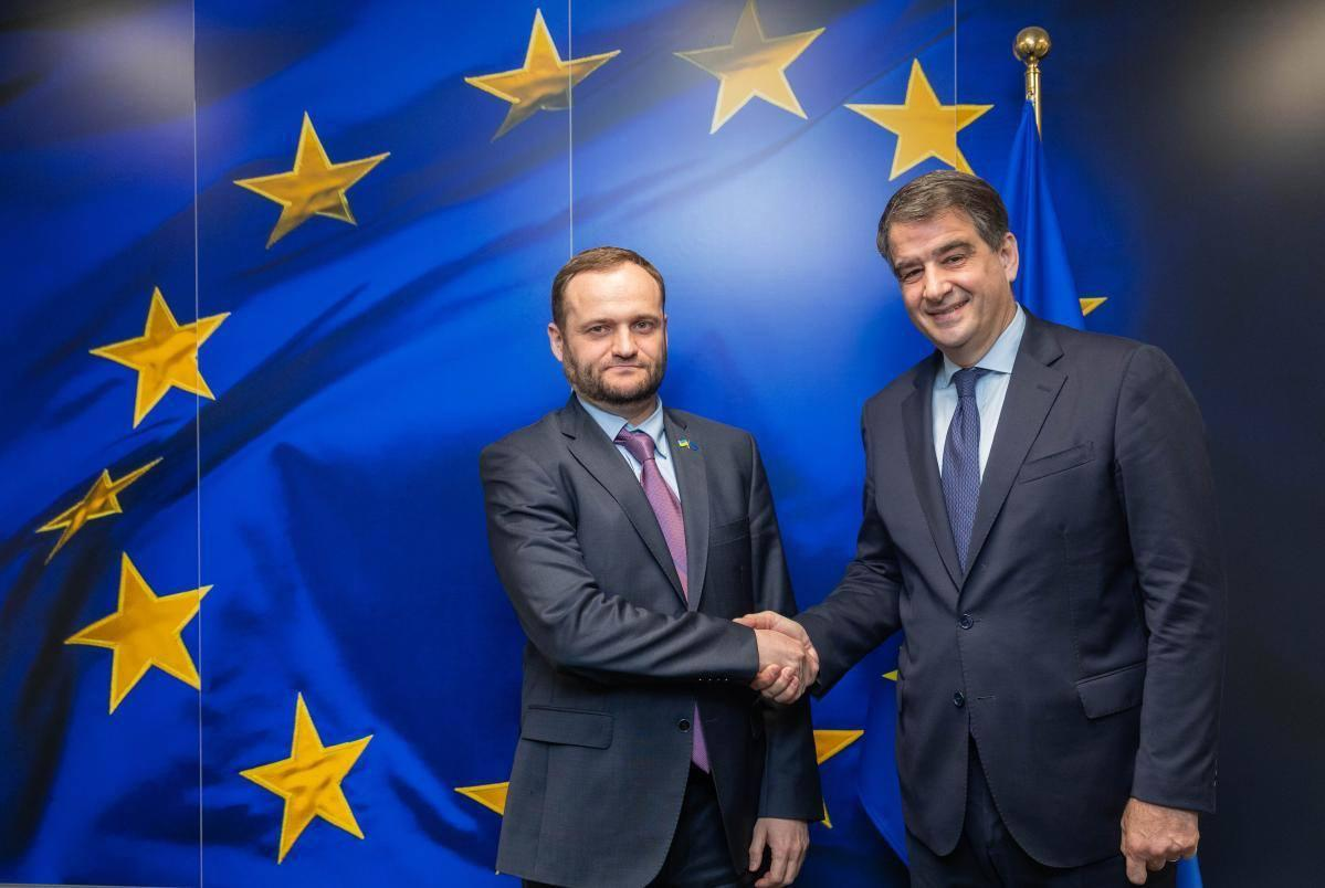 Двоє чоловіків у ділових костюмах тиснуть руки на фоні великого прапора Європейського Союзу. Один із них має значок із прапором України на лацкані піджака.