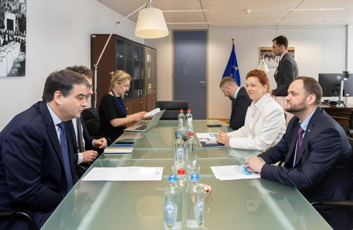 Група людей у діловому вбранні проводить офіційну зустріч за довгим скляним столом у кабінеті на фоні прапора ЄС.