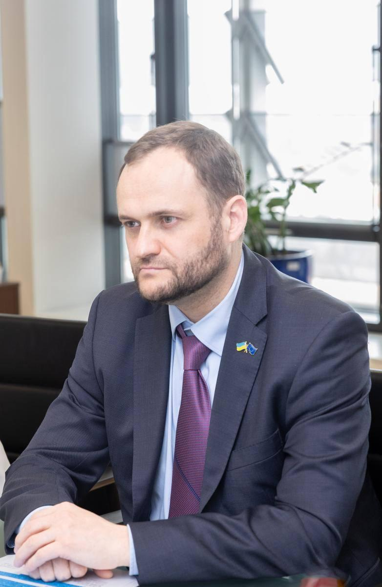 Чоловік у діловому костюмі та краватці сидить за столом у світлому офісі. На лацкані його піджака — значок із прапорами України та Європейського Союзу.