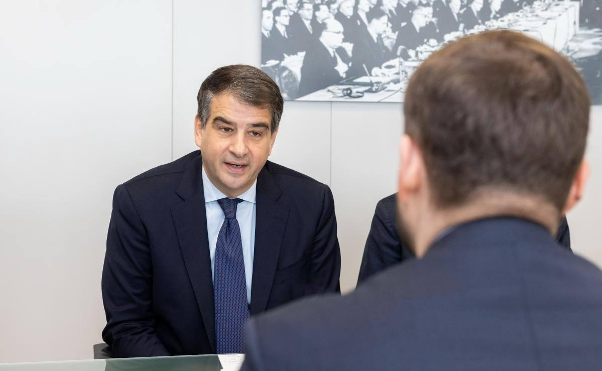 Чоловік у діловому костюмі та краватці спілкується зі співрозмовником за столом у кабінеті. На стіні позаду нього висить чорно-біле фото офіційного заходу.