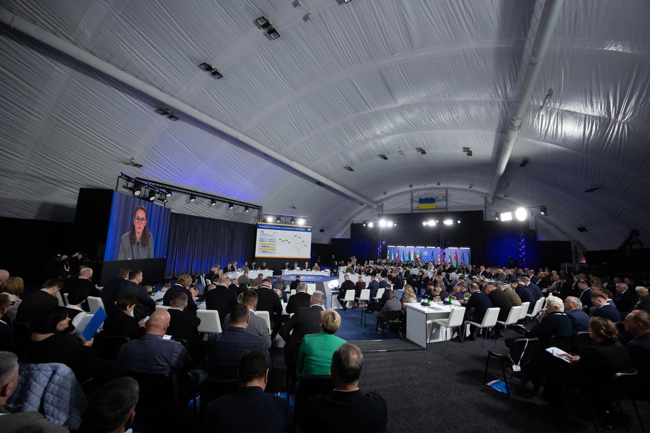 Великий зал під арковим накриттям, заповнений учасниками масштабної конференції. Люди сидять за столами та в рядах перед великими екранами, на яких транслюється виступ. У глибині залу — стійка з міжнародними прапорами.