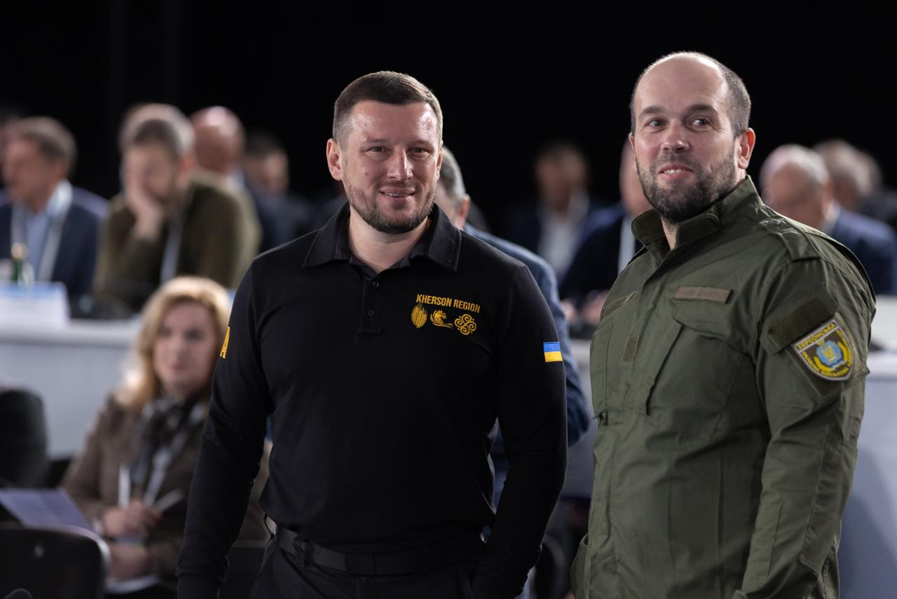 Двоє чоловіків у чорній тенісці з написом «KHERSON REGION» та зеленій тактичній куртці стоять у залі на фоні численних учасників офіційного заходу.