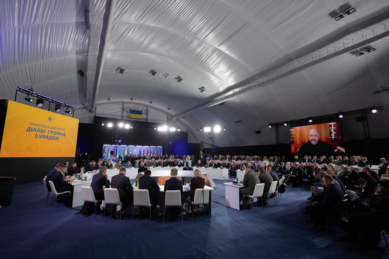 Масштабний захід у великому залі під арковим накриттям. Учасники сидять за білими столами, розставленими колом та рядами. На великих екранах транслюється назва події «Діалог громад з Урядом» та відеозв'язок зі спікером. На задньому плані розміщено низку державних прапорів.