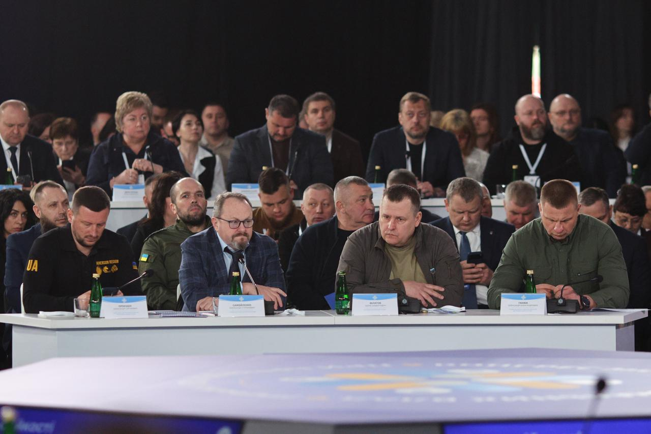 Численна група учасників офіційного заходу сидить у залі. У першому ряду за довгим білим столом четверо чоловіків перед мікрофонами та іменними табличками, за ними — кілька рядів слухачів. На передньому плані видно частину круглого столу з логотипом.