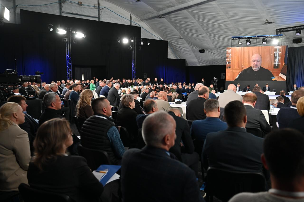 Вид із залу на масштабний форум у великому приміщенні під тентовим дахом. Численна аудиторія сидить у кріслах, спостерігаючи за виступом спікера на великому екрані над сценою. У залі встановлено професійне освітлення та відеокамери.