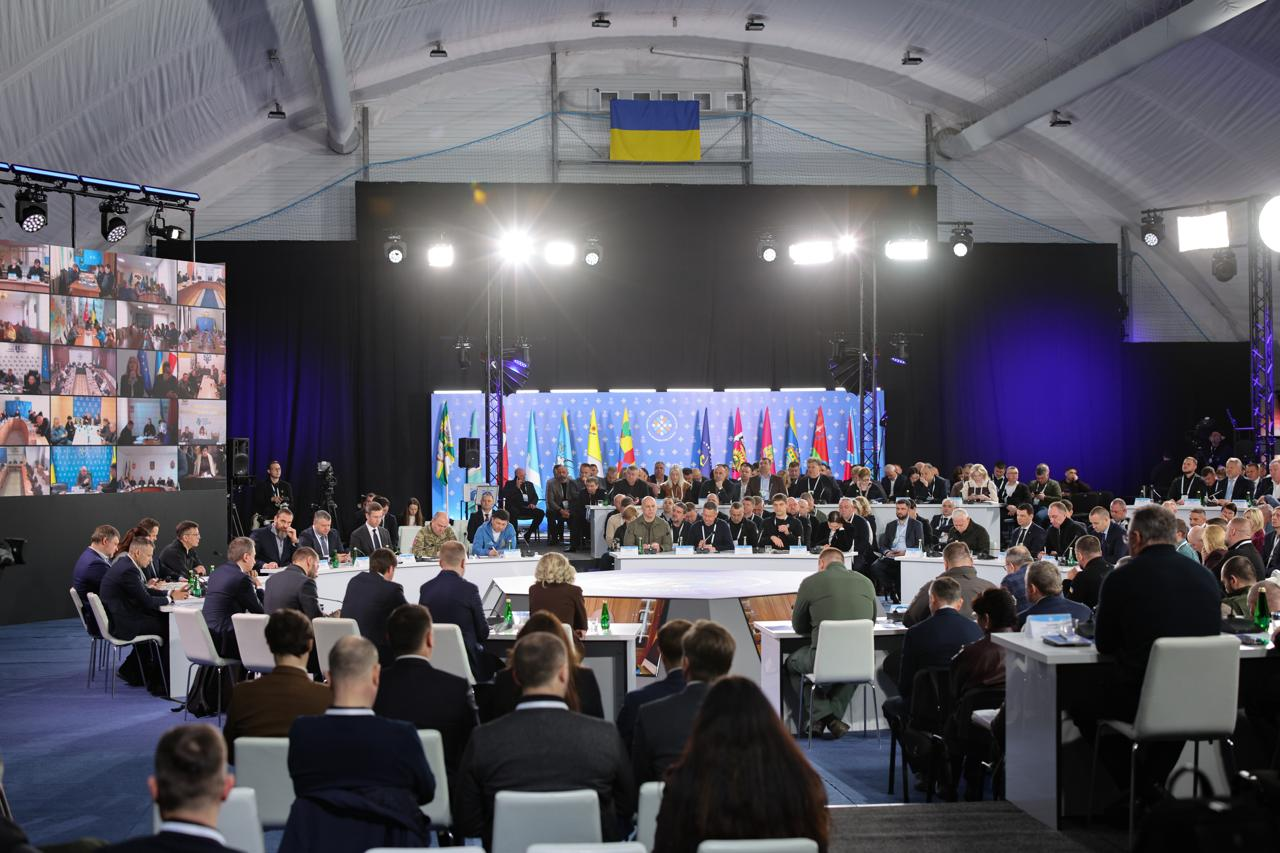 Великий зал під арковим накриттям під час офіційного форуму. У центрі — круглий стіл, за яким сидять учасники, оточений численною аудиторією в залі. На задньому плані — сцена з прапорами та яскравим освітленням, а зліва — великий екран із багатьма вікнами відеозв'язку. Вгорі під стелею закріплений прапор України.