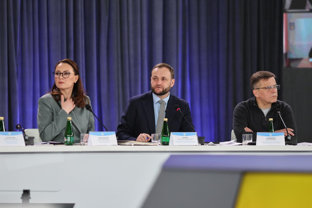 Троє людей сидять за довгим білим столом із мікрофонами та пляшками води під час офіційної зустрічі на тлі синіх штор. Перед ними розміщені іменні таблички.