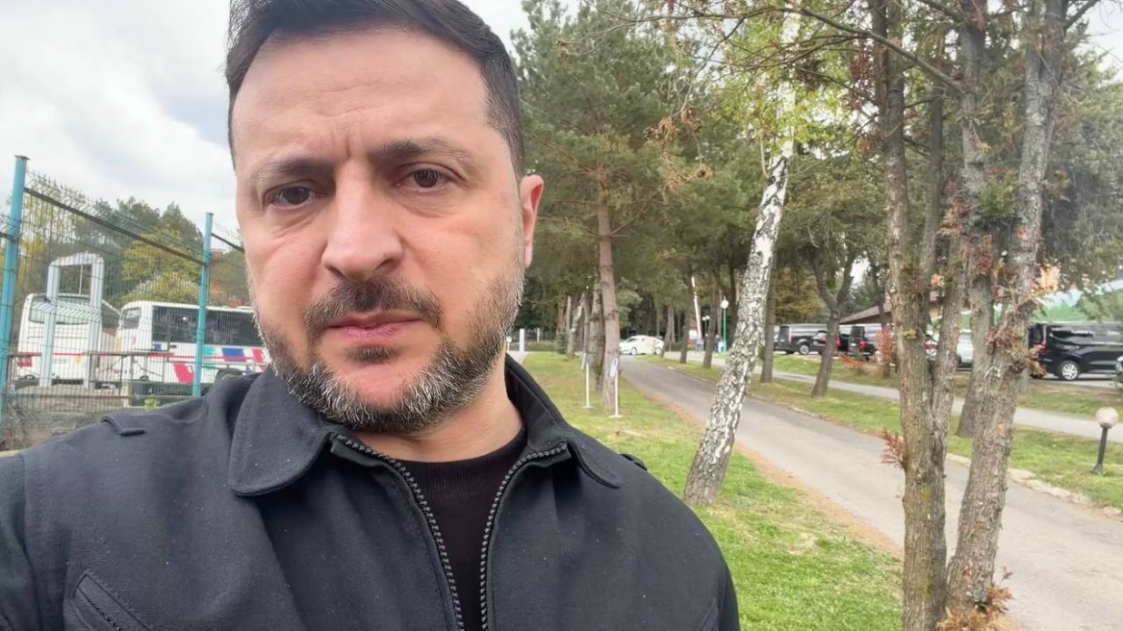 Чоловік у чорній куртці на фоні паркової зони з деревами, дорогою та припаркованими автомобілями й автобусами за парканом.