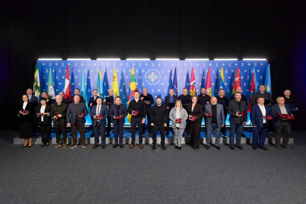 Групова фотографія понад двадцяти осіб, які стоять у два ряди на сцені. Учасники тримають у руках невеликі нагороди або футляри. Тло оформлене синім банером із символікою та численними прапорами областей і міст України.