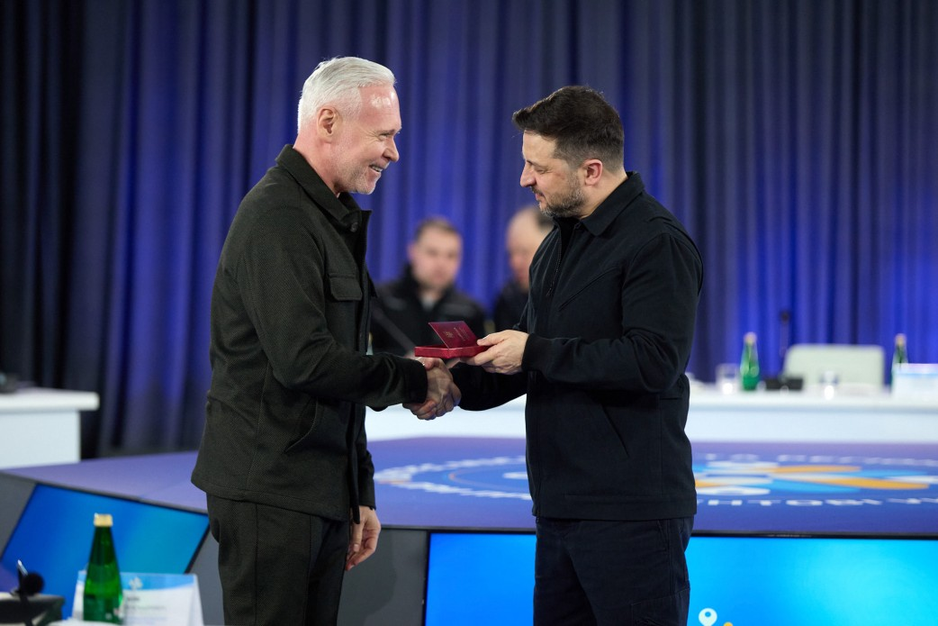 Двоє чоловіків потискають руки під час урочистого заходу. Один із них передає іншому відкритий футляр із державною нагородою та посвідченням. Дія відбувається на фоні сцени з логотипом форуму та синіх штор.