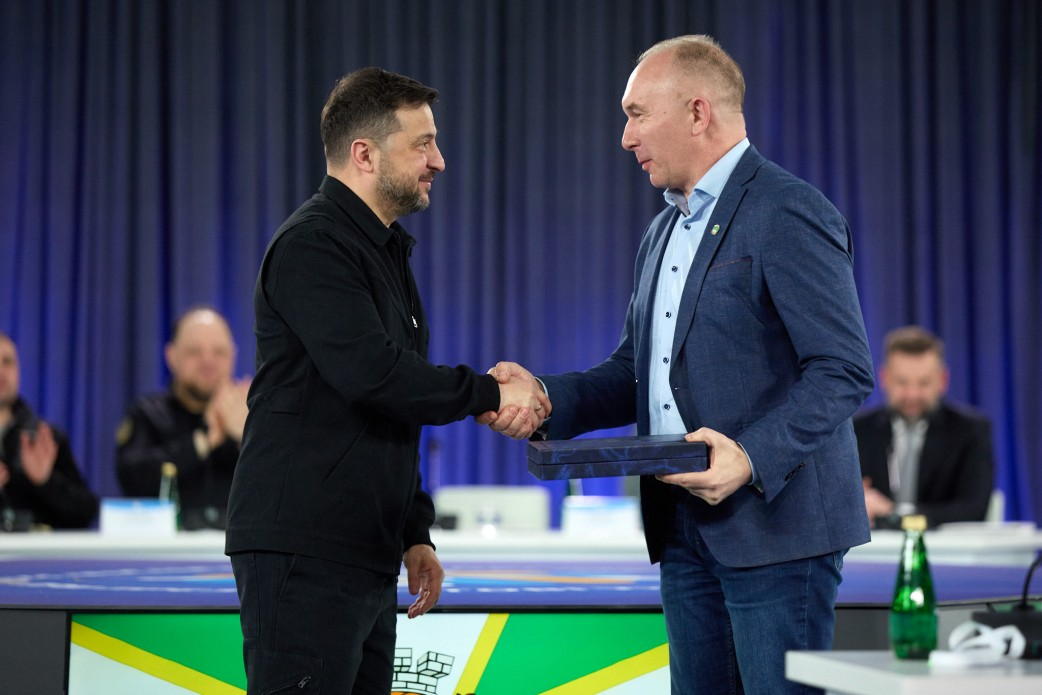 Двоє чоловіків потискають руки під час офіційної церемонії. Один із них тримає синій футляр із нагородою. На задньому плані — сині штори та присутні за столом учасники заходу.