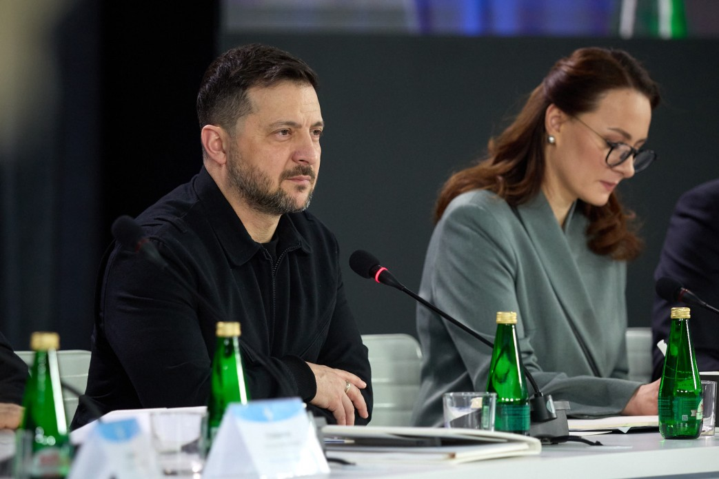 Чоловік у чорному одязі та жінка в сірому піджаку й окулярах сидять за білим робочим столом під час офіційного засідання. Перед ними встановлені мікрофони, стоять пляшки з водою та розкладені папери.