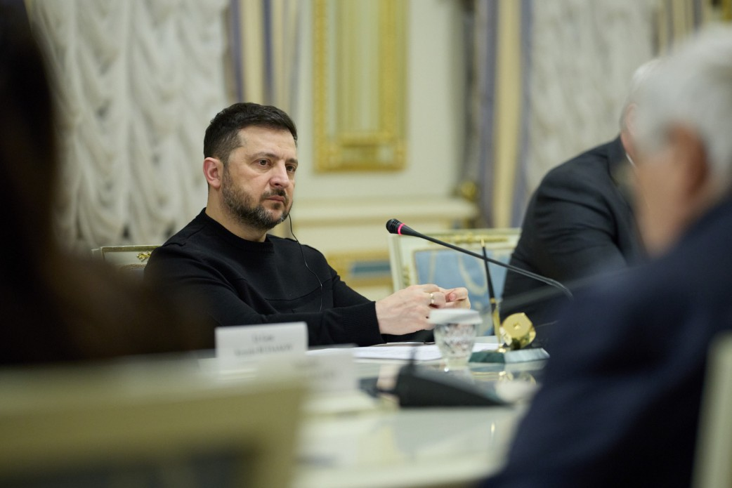 Чоловік у чорному одязі зосереджено слухає співрозмовника під час офіційної зустрічі. Він сидить за столом з мікрофоном у світлому залі з класичним інтер'єром.