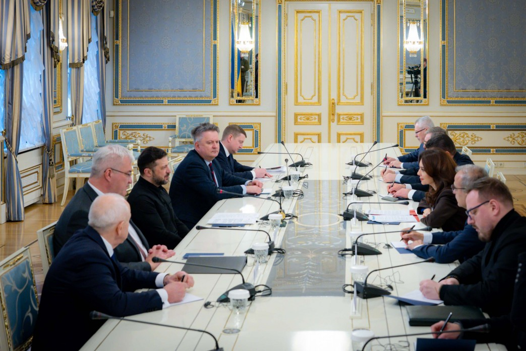 Офіційна зустріч двох делегацій за довгим столом у світлій залі з класичним декором. Учасники в діловому одязі ведуть переговори перед мікрофонами, тримаючи ручки та папери.