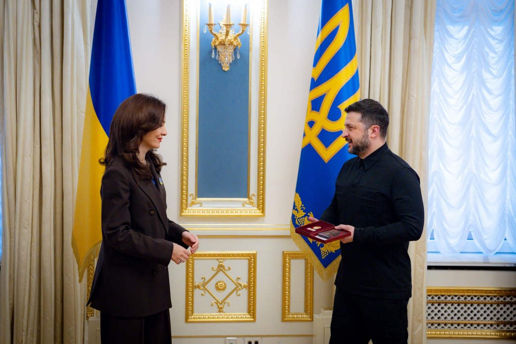 Офіційна зустріч двох осіб у світлій залі на фоні державних прапорів. Чоловік у темному одязі вручає жінці в діловому костюмі нагороду у відкритому футлярі.