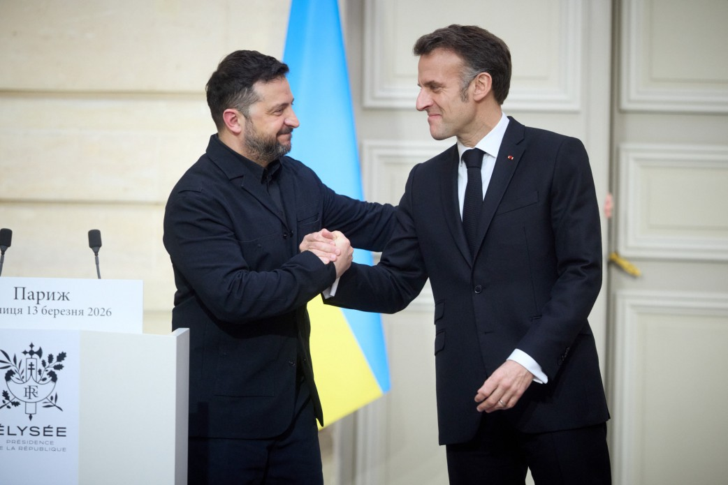 Офіційне привітання двох політичних лідерів у Парижі. Чоловіки усміхаються та потискають один одному руки на фоні державного прапора біля трибуни з емблемою Єлисейського палацу.
