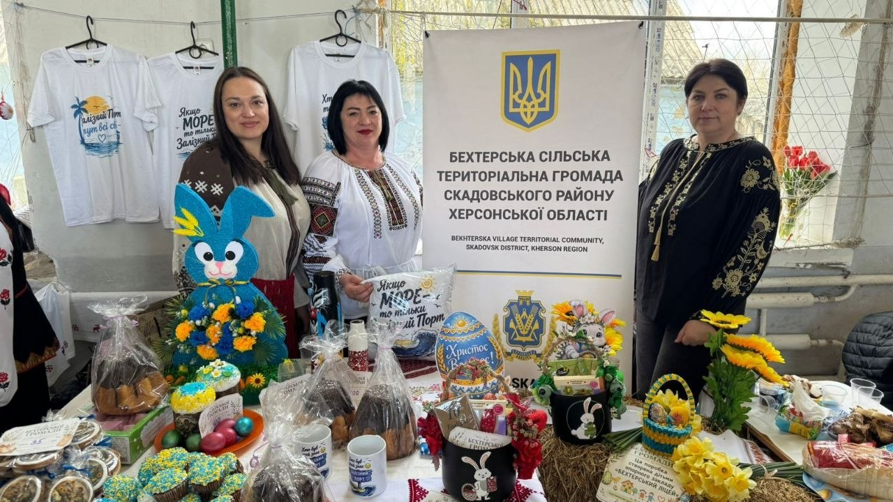 Три жінки у вишиванках представляють стенд територіальної громади на благодійному ярмарку. Стіл заставлений великодніми пасками, крашанками та сувенірами, а на фоні розміщено інформаційний банер із гербом України та розвішано футболки з принтами.