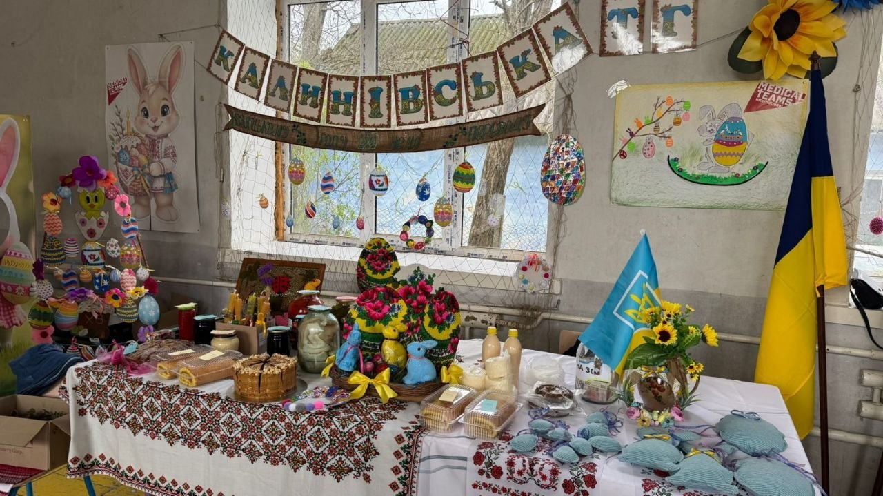 Благодійний ярмарок у приміщенні, прикрашеному до Великодня українською символікою та дитячими малюнками. На столі, застеленому вишитою скатертиною, представлені паски, солодощі, крашанки та вироби ручної роботи.