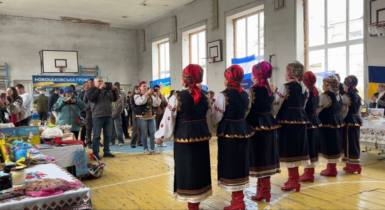 Виступ фольклорного колективу в українських народних костюмах посеред спортивного залу під час благодійного ярмарку. Глядачі знімають дійство на телефони, а вздовж стін розміщено столи з продукцією та патріотичною символікою громад.