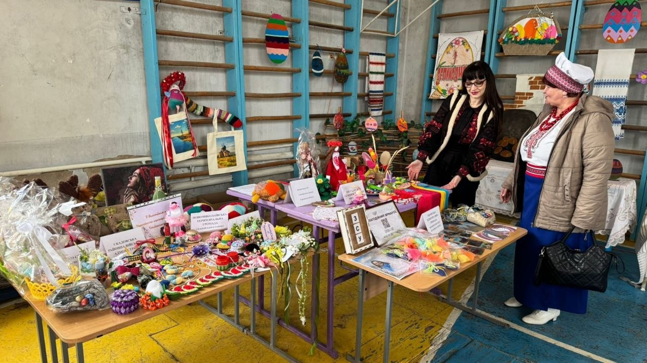 Дві жінки в українському вбранні розглядають вироби ручної роботи на благодійному ярмарку. На столах представлені ляльки-мотанки, прикраси, сумки та великодній декор, а на фоні шведської стінки розміщено вишиті рушники та тематичні інсталяції.