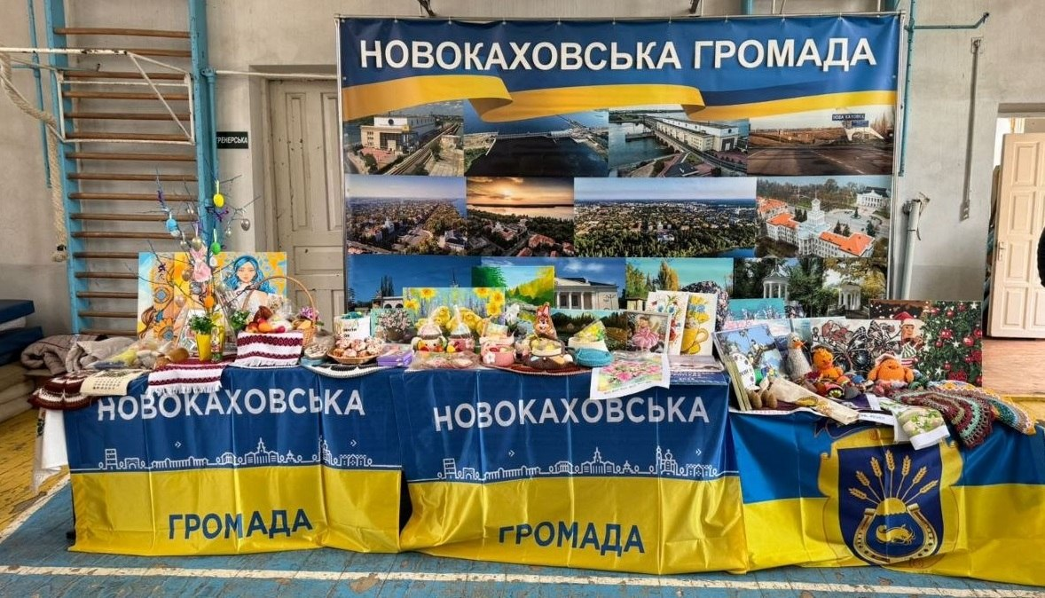 Презентаційний стенд Новокаховської громади на офіційному заході. Стіл із сувенірами, картинами та великоднім декором застелений синьо-жовтими скатертинами на фоні великого інформаційного банера з краєвидами міста.