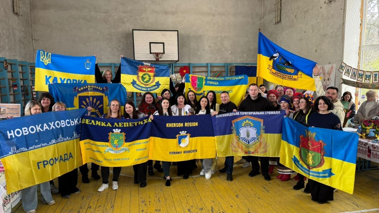 Група людей у спортивній залі тримає прапори територіальних громад Херсонщини під час офіційного заходу. На передньому плані видно великі синьо-жовті стяги з назвами та гербами громад, зокрема Новокаховської, Великолепетиської, Зеленопідської та інших.