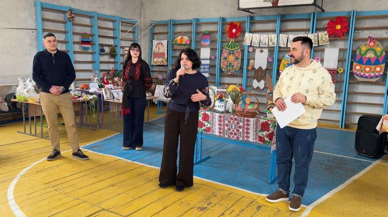 Виступ організаторів або гостей під час благодійного заходу в спортивному залі. Жінка з мікрофоном звертається до присутніх, поруч стоять інші учасники події на фоні шведської стінки, прикрашеної вишитими рушниками та великоднім декором.