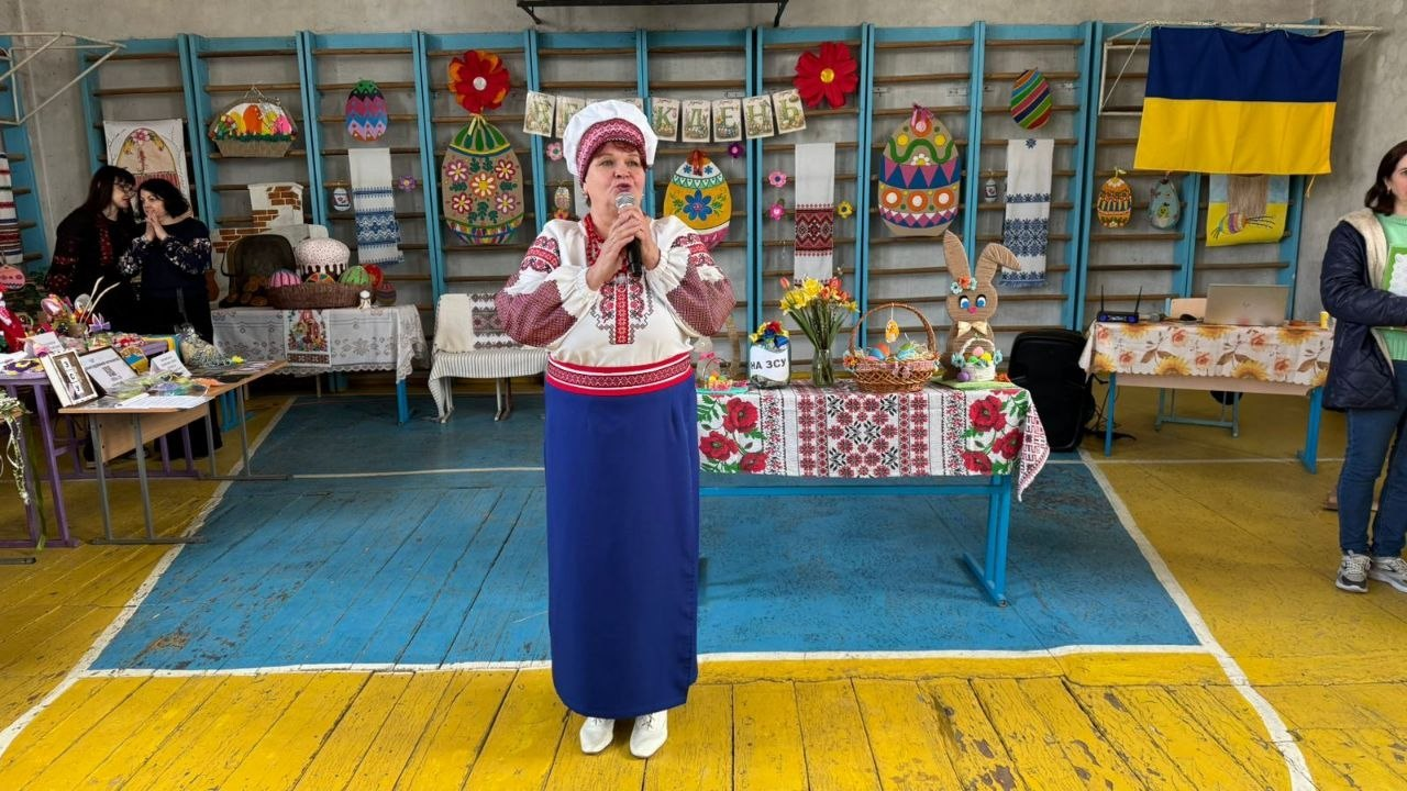 Жінка в українському народному строї виступає з мікрофоном у спортивній залі, прикрашеній до Великодня. На фоні шведської стінки розміщено святкові декорації, вишиті рушники та державний прапор України, а поруч на столах представлені пасхальні кошики та квіти.