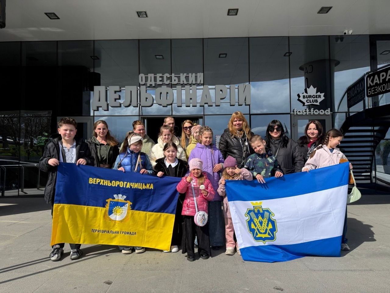 Група дорослих та дітей позирує для спільного фото перед будівлею Одеського дельфінарію. Вони тримають розгорнуті прапори: ліворуч — прапор Верхньорогачицької територіальної громади, праворуч — біло-синій стяг із гербом Херсонської області.