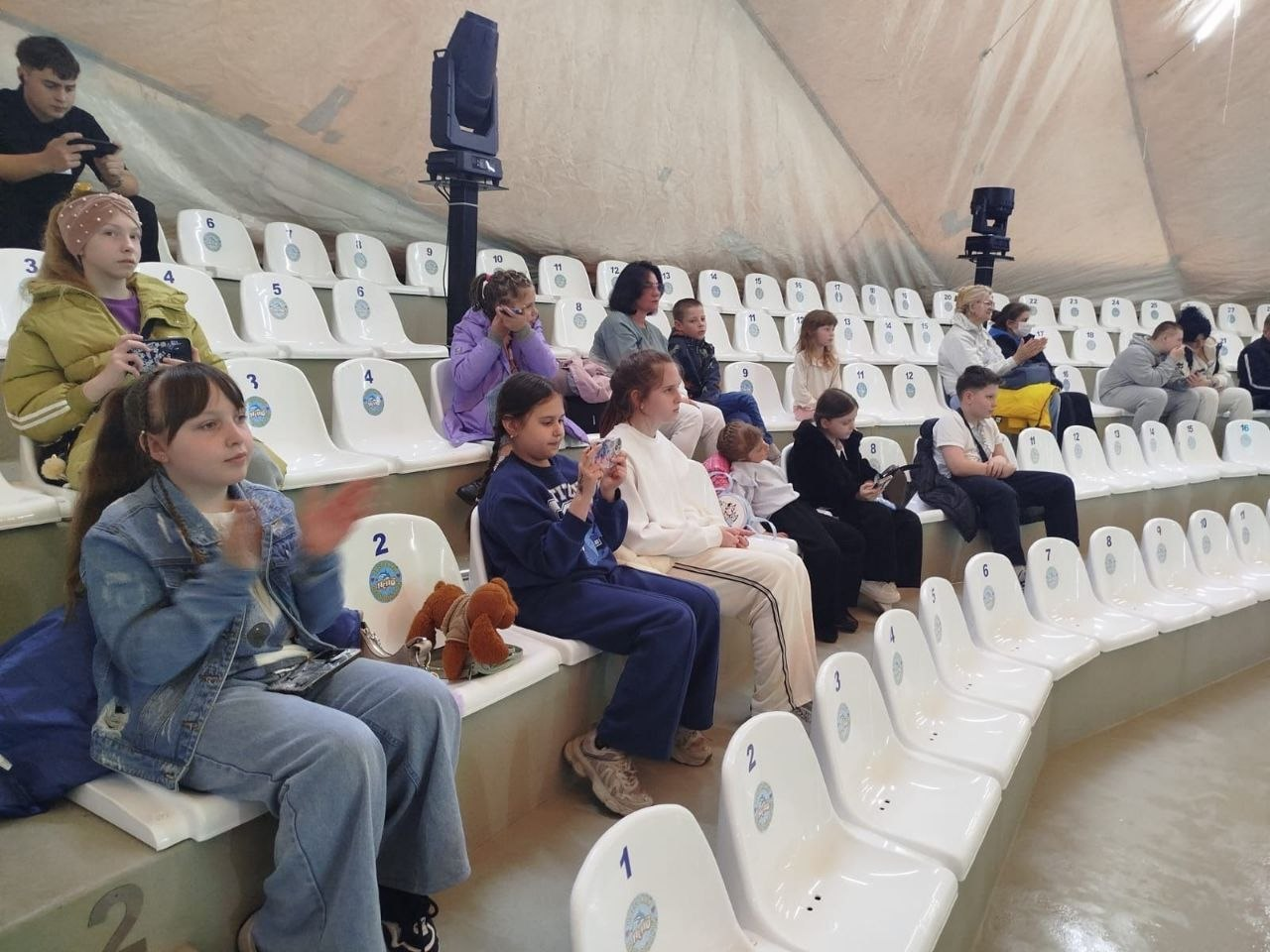 Група дітей та дорослих сидить на трибунах дельфінарію під час вистави. Глядачі зосереджено спостерігають за дійством, дехто знімає подію на телефони або аплодує, сидячи на білих пластикових кріслах.