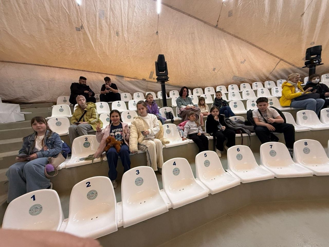 Глядачі різного віку розмістилися на білих пластикових сидіннях у залі під куполом. Вони чекають на початок вистави або відпочивають у перерві, дехто користується мобільними телефонами.