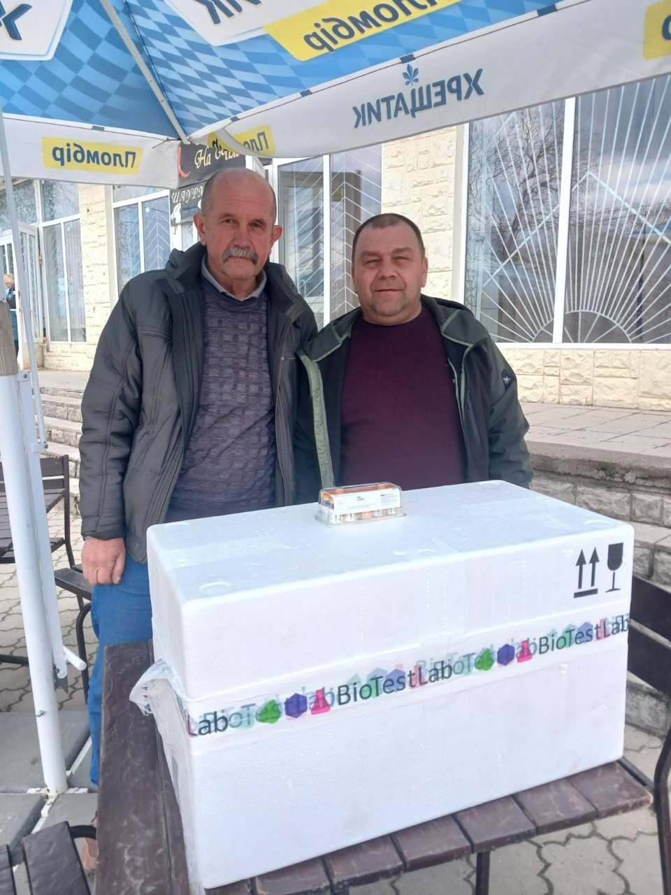 Двоє чоловіків стоять біля великого білого термобокса з маркуванням «BioTestLab», на якому лежить маленька упаковка з флаконами. Зустріч відбувається на вулиці під брендованою парасолькою на фоні будівлі.