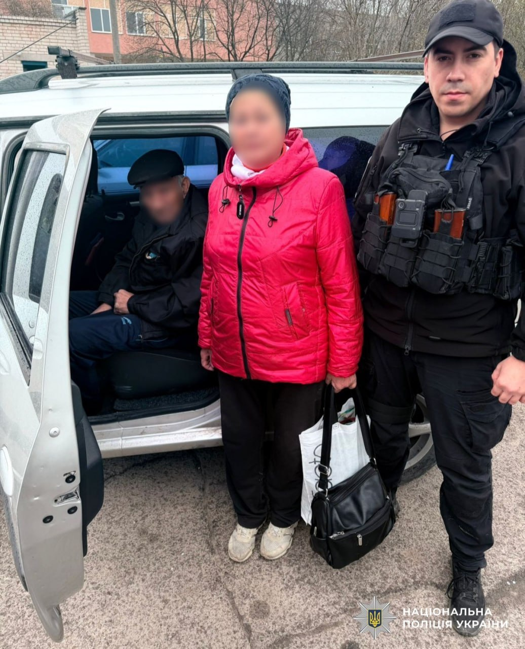 Правоохоронець у тактичному спорядженні стоїть поруч із двома цивільними особами біля відчинених дверей автомобіля. У нижньому куті розміщено логотип Національної поліції України.