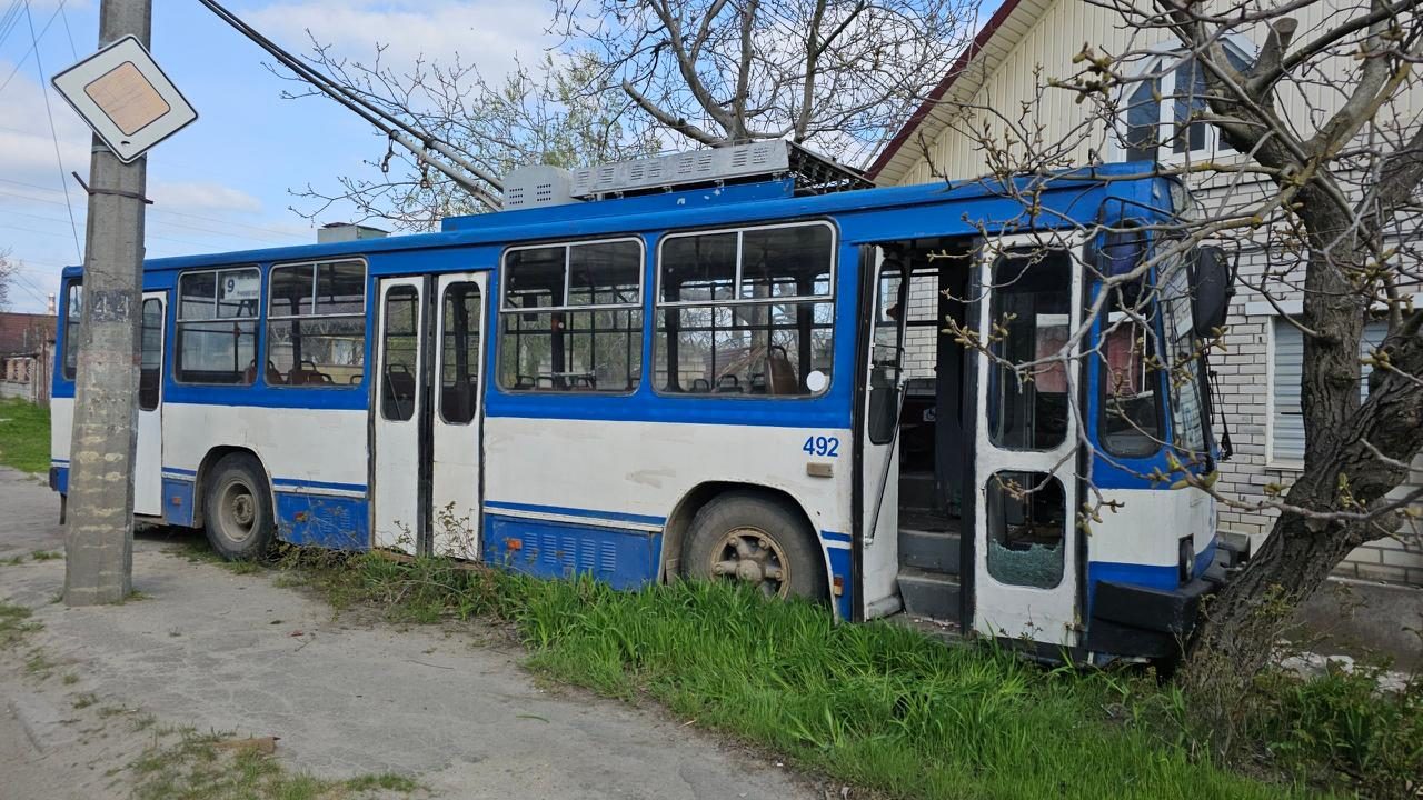 На фото зображений покинутий синьо-білий тролейбус ЮМЗ Т2 з бортовим номером 492, що стоїть на узбіччі дороги в траві біля дерев та житлового будинку. Вікна транспортного засобу пошкоджені, а передні двері відчинені.
