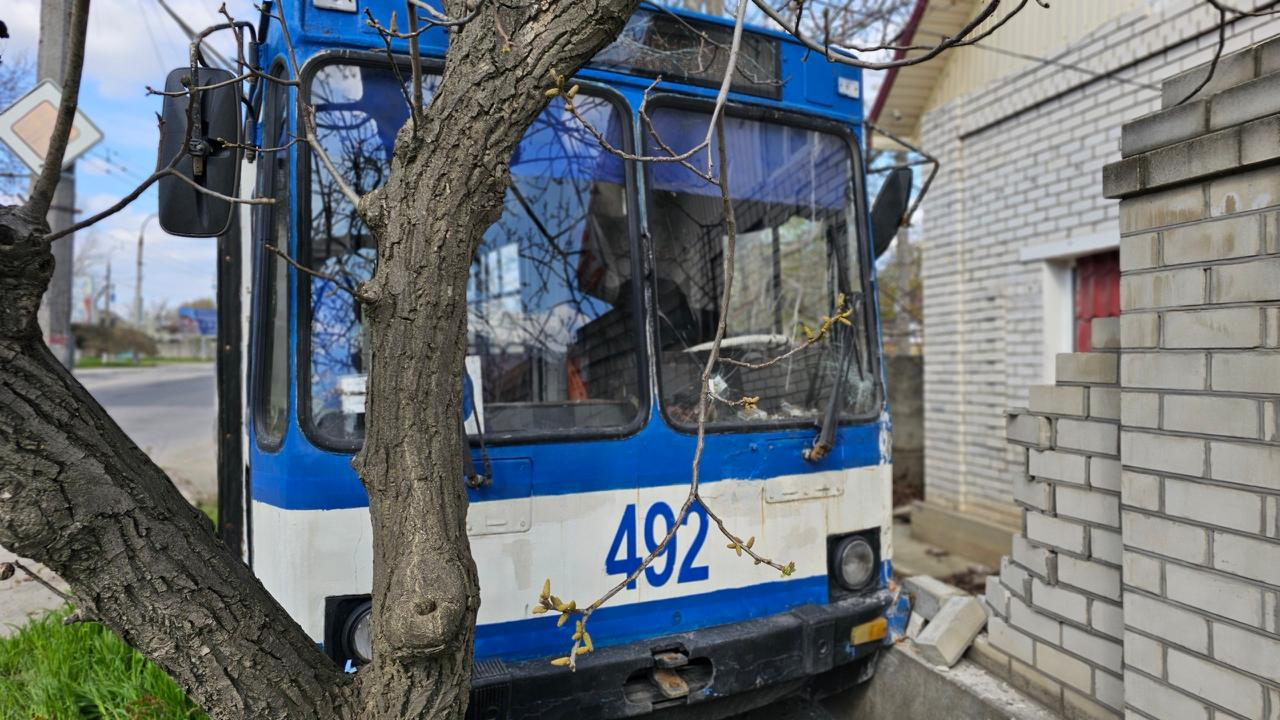 На фото великим планом зображена передня частина покинутого синьо-білого тролейбуса ЮМЗ Т2 з бортовим номером 492. Транспортний засіб стоїть впритул до дерева та цегляного паркану, лобове скло має тріщини.