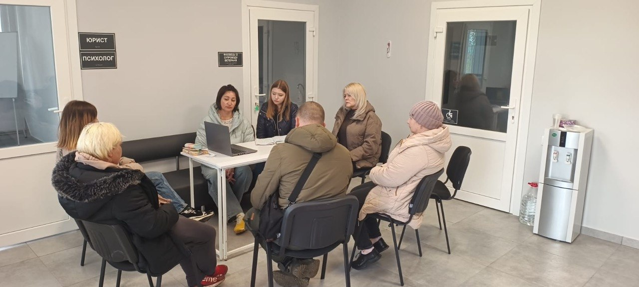 Група людей сидить за столом та вздовж стін у світлому офісному приміщенні, проводячи зустріч або консультацію. На стіні помітні таблички «Юрист» та «Психолог», а поруч стоїть кулер для води.