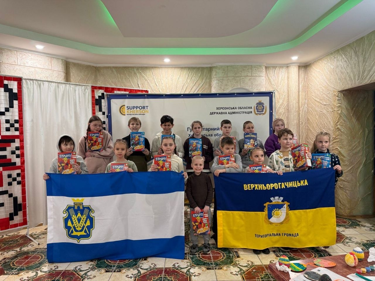 Група дітей із гуманітарною допомогою в руках тримає прапори Херсона та Верхньорогачицької громади на фоні офіційних банерів.