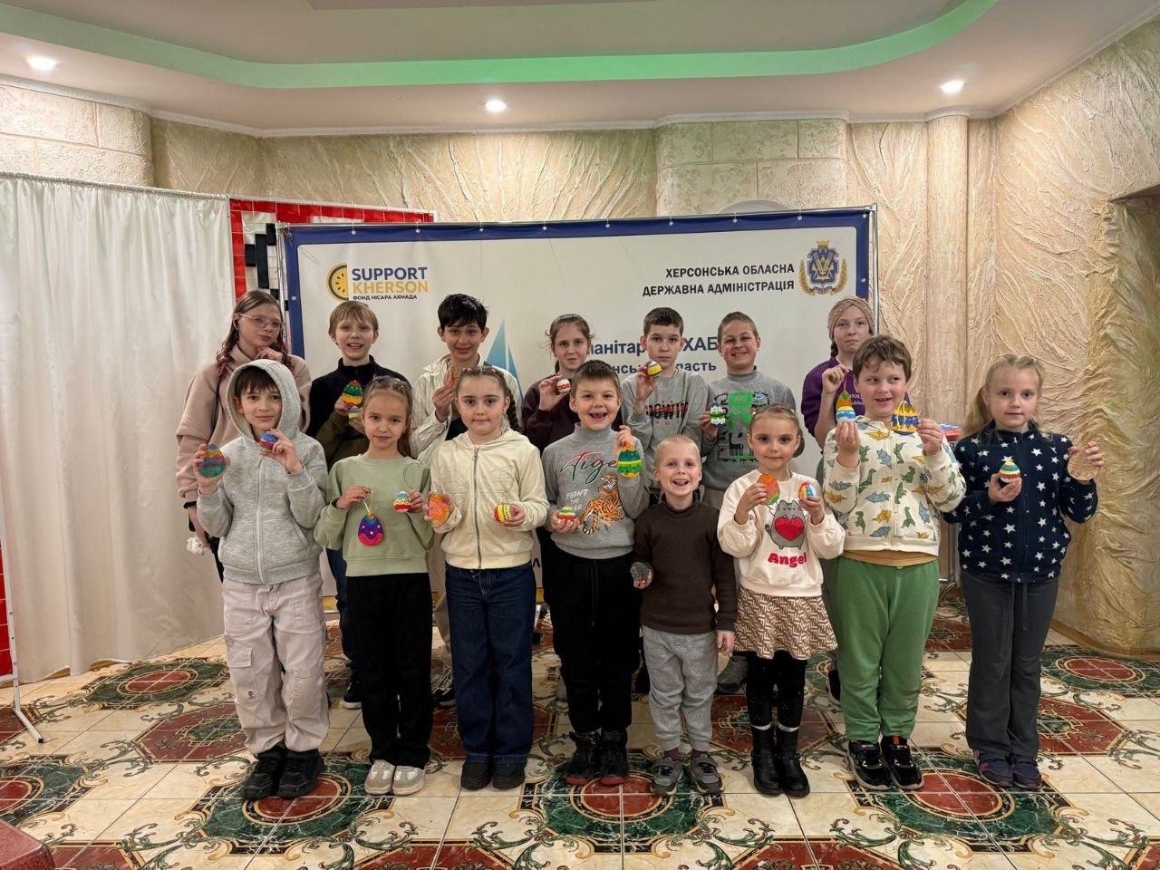 Група дітей стоїть у приміщенні на фоні банера Херсонської ОВА та тримає в руках розфарбовані великодні писанки.