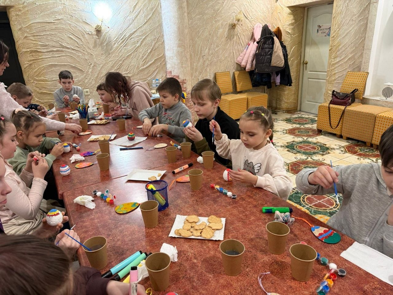 Діти сидять за довгим столом у приміщенні та розфарбовують великодні писанки фарбами й маркерами. На столі також стоять стаканчики з водою та печиво.