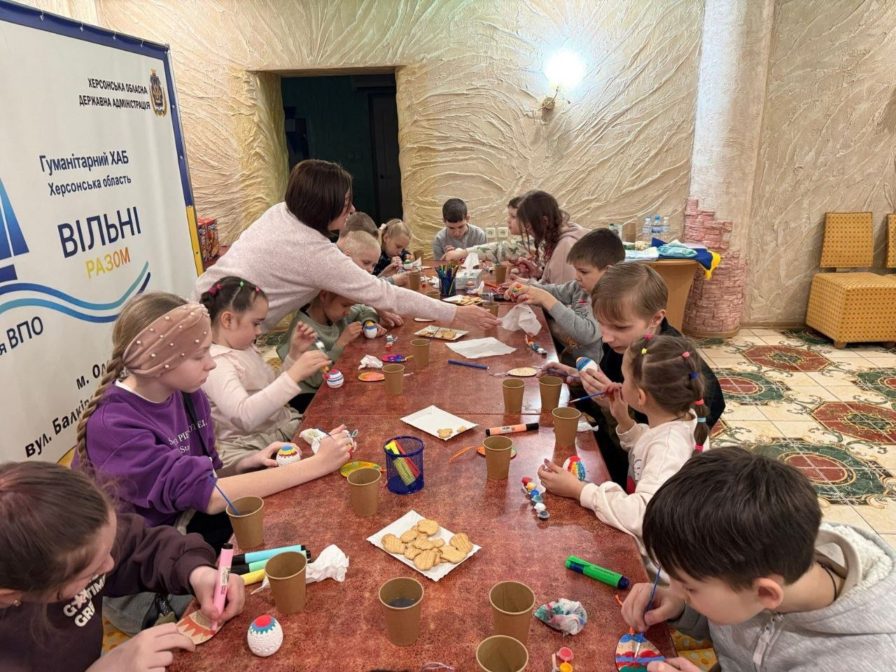 Діти та дорослі разом розписують великодні яйця за довгим столом у гуманітарному хабі. Поруч стоїть інформаційний банер Херсонської ОВА «Вільні разом».