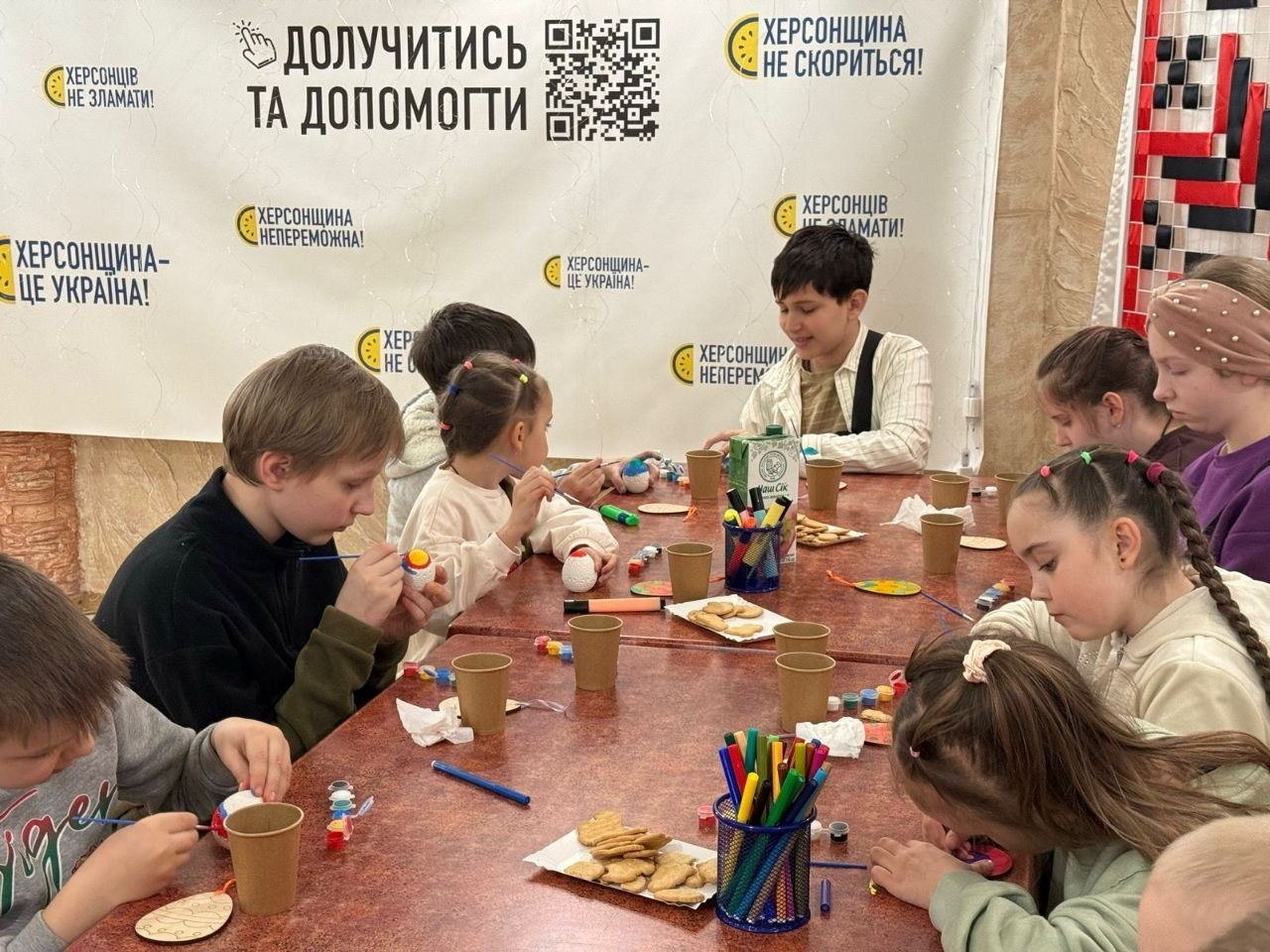 Діти займаються творчістю за столом на фоні банера з патріотичними гаслами та QR-кодом для допомоги. Вони розписують великодні яйця, використовуючи фарби, пензлики та маркери.
