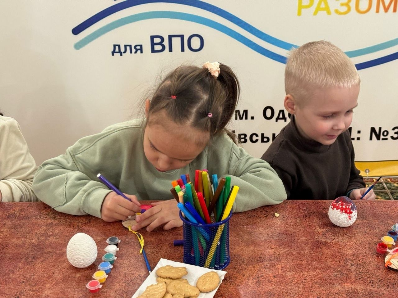 Двоє дітей захоплено розписують великодні яйця за столом, на якому лежать фарби, маркери та печиво. За ними видніється частина банера з написом «для ВПО».