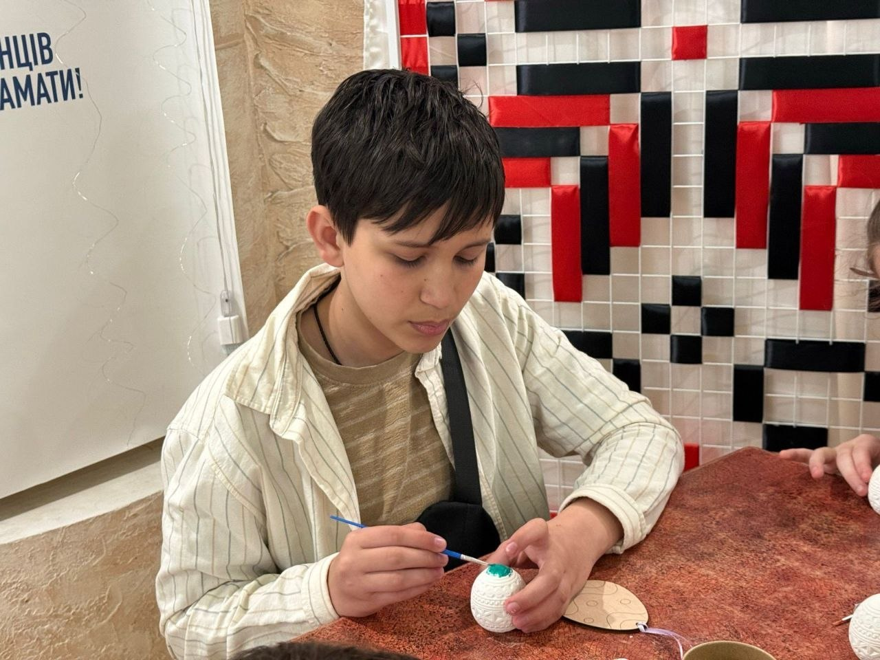 Підліток зосереджено розмальовує великоднє яйце пензликом, сидячи за столом на фоні декоративного панно з червоно-чорним орнаментом.